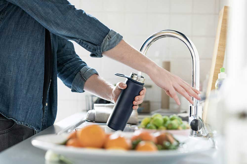 Filling water bottle at the kitchen sink, with fruit and vegatables around